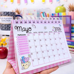 Puede incluir: Un calendario de escritorio a cuadros rosa y blanco para mayo de 2026, con encuadernación en espiral y una lista de prioridades. El calendario tiene una linda ilustración de una taza con lápices y tijeras.