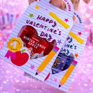 Könnte beinhalten: Zwei Valentinstags-Geschenkanhänger von Bath & Body Works. Die weißen Anhänger mit blauen Linien tragen den Text "Happy Valentine's Day". Einer hat ein rotes PocketBac in Apfelform, der andere ein lila PocketBac. Gelbe Bleistifte und Sterne verzieren die Anhänger.