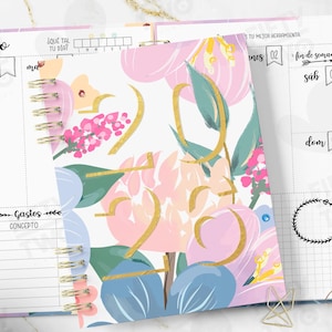 May include: A floral-patterned planner with a gold spiral binding. The cover displays a watercolour design with pink, blue, and green flowers, and gold accents. The planner is open to a page with the word "enero" and other Spanish text.