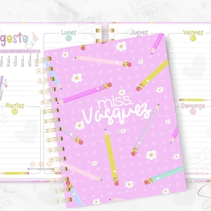 May include: A spiral-bound planner with a pink cover featuring the text "miss Vasquez" and colorful pencils. The planner is open to show a weekly layout with the month of August and the days of the week in Spanish.