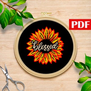 May include: Cross stitch pattern featuring a sunflower with the word "Blessed" in the center. The sunflower is yellow and orange with a black background.