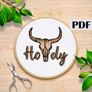 Könnte beinhalten: Ein weißer Stickrahmen mit einem braunen Stierkopf und dem Text "Howdy" in brauner Kursivschrift. Der Rahmen befindet sich auf einer Holzoberfläche.