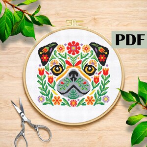 以下が含まれることがあります： 木製の刺繍枠の中に、カラフルな花の模様で囲まれたパグ犬の顔のクロスステッチパターン。デザインは赤、オレンジ、黄色、緑の花が特徴です。「PDF」の文字が右上隅にあります。