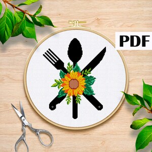 Può includere: Schema di punto croce con un girasole e una silhouette nera di una forchetta, un coltello e un cucchiaio. Lo schema è in un cerchio da ricamo bianco.