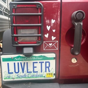May include: A red Jeep with a license plate that reads "LUVLETR South Carolina" and the text "Nothing Could Be Finer" above it. The license plate has a February 24th date sticker. There are white heart stickers on the back of the Jeep.
