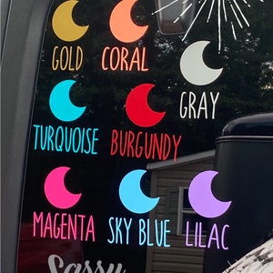May include: A black car window with a variety of moon-shaped decals in different colors, including gold, coral, gray, turquoise, burgundy, magenta, sky blue, and lilac. The decals are arranged in rows and are labeled with their corresponding colors. The text "NEW COLORS!" is also visible on the window. The text "Sassy Decal Co. AN ETSY SHOP" is visible at the bottom of the window.
