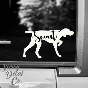May include: Black and white silhouette of a dog with the word "Scout" written on its back. The silhouette is on a car window.