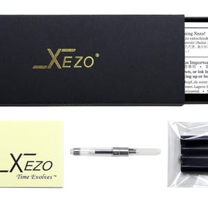 Pode incluir: Uma caixa preta com letras douradas que dizem "_XEZO" e um cart&atilde;o amarelo a condizer com o mesmo log&oacute;tipo e o texto "Time Evolves". Tamb&eacute;m est&atilde;o inclu&iacute;dos uma caneta transparente, um pacote de cartuchos de tinta preta e um papel com texto.