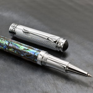 May include: A silver and abalone shell rollerball pen with a textured silver barrel and a black gemstone accent. The pen is lying on a dark gray surface.