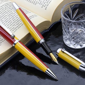 May include: Two writing instruments featuring a yellow and red barrel design with silver-toned accents. One is a fountain pen with a gold-colored nib and a separate yellow cap, while the other is a ballpoint pen with a silver tip and a clip.