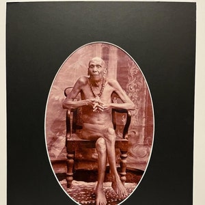May include: A black and white photograph of an elderly person sitting on a chair. The person is wearing a beaded necklace and has a thin build. The photograph is framed in a black oval mat.