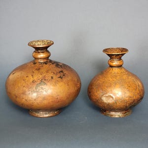 Puede incluir: Dos jarrones antiguos de cobre con intrincados diseños grabados. El jarrón más grande tiene una base más ancha y una forma más redondeada, mientras que el jarrón más pequeño tiene una base más estrecha y una forma más alargada.