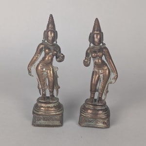 Puede incluir: Dos estatuas de bronce de figuras femeninas. Cada estatua tiene un tocado cónico, rasgos faciales detallados y posa con un brazo levantado. Las figuras se alzan sobre una base decorativa. Las estatuas se exhiben sobre un fondo liso.