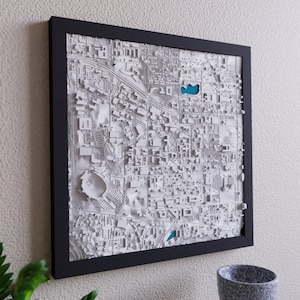 May include: A white 3D printed map of a city in a black frame. The map is raised and textured, showing the city's streets and buildings.