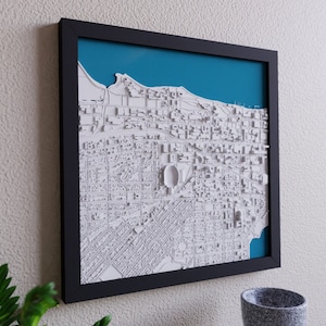 K&ouml;nnte beinhalten: Eine gerahmte 3D-Karte einer Stadt mit wei&szlig;en Geb&auml;uden und einem blauen Wassermerkmal. Die Karte ist in Schwarz gerahmt und h&auml;ngt an einer wei&szlig;en Wand.