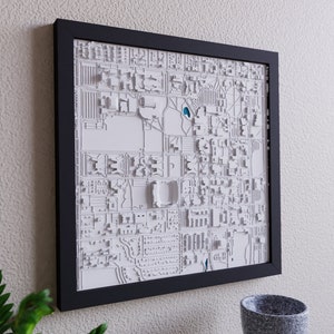 May include: A white 3D relief map of a city in a black frame. The map is raised and textured, showing the buildings and streets of the city. The map is mounted on a white wall.