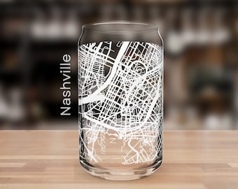 May include: A clear glass can-shaped drinking glass with a white map of Nashville. The word "Nashville" is printed vertically on the side of the glass. The glass sits on a wooden surface with a blurred background.