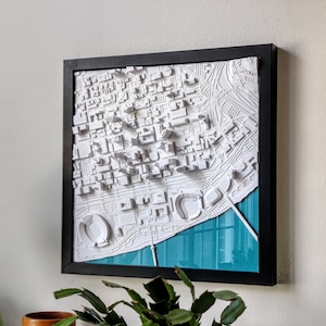 May include: A framed 3D map of a city with white buildings and a blue water feature. The map is framed in black and has a white mat.