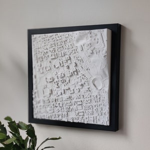 May include: A white 3D map of a city, framed in black. The map shows detailed building and street layouts. The artwork is mounted on a white surface and is displayed on a white wall. A potted plant and a brown mug are in the foreground.