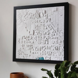 May include: A white 3D printed map of a city in a black frame. The map is raised and textured, showing the buildings and streets of the city. The map is hanging on a white wall.