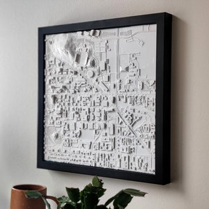 May include: A framed, three-dimensional white map with a black frame. The map depicts a detailed urban landscape, showcasing buildings, roads, and other features. The artwork is mounted on a neutral-colored wall, suggesting a modern interior design.
