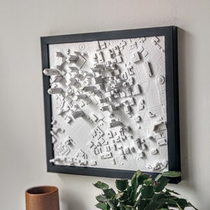 May include: A white 3D cityscape model in a black frame. The model is raised and textured, giving it a realistic look. The cityscape is a detailed representation of a city with buildings, streets, and parks.