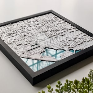 May include: A framed 3D printed map of a city, with white buildings and a blue water feature. The map is set against a black background and framed in a black frame.