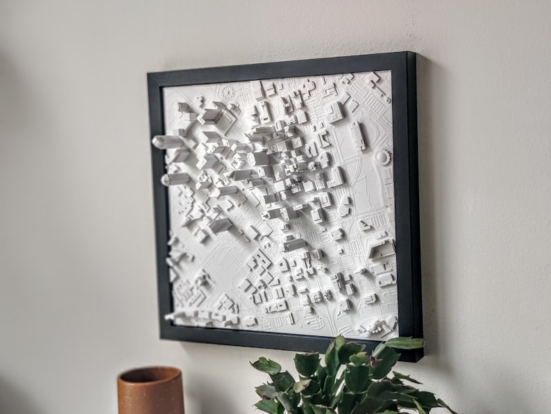 May include: A white 3D cityscape model in a black frame. The model is raised and textured, giving it a realistic look. The cityscape is a detailed representation of a city with buildings, streets, and parks.