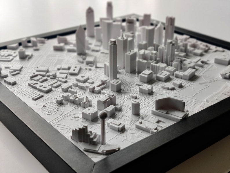 May include: A white 3D printed model of a city skyline, featuring tall buildings and a detailed street layout. The model is framed in black and sits on a white surface.
