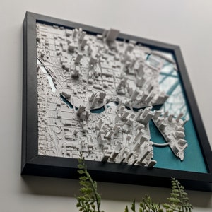 Puede incluir: Un mapa de ciudad en 3D enmarcado con edificios blancos y una caracter&iacute;stica de agua azul turquesa. El mapa est&aacute; enmarcado en un marco negro.