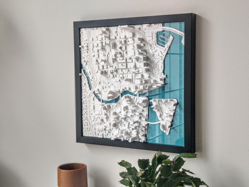 Puede incluir: Un mapa 3D enmarcado de una ciudad con edificios blancos y una caracter&iacute;stica de agua azul. El mapa est&aacute; sobre un fondo azul verdoso.
