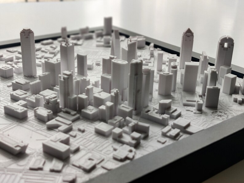 May include: A white 3D printed model of a city skyline, featuring tall buildings and streets. The model is displayed in a black frame.
