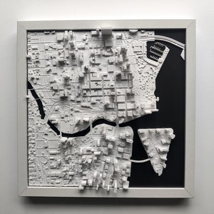 Puede incluir: Un modelo impreso en 3D de un horizonte de ciudad, en blanco, enmarcado en blanco y sobre un fondo negro. El modelo presenta edificios y calles detalladas, con un r&iacute;o que serpentea por la ciudad.