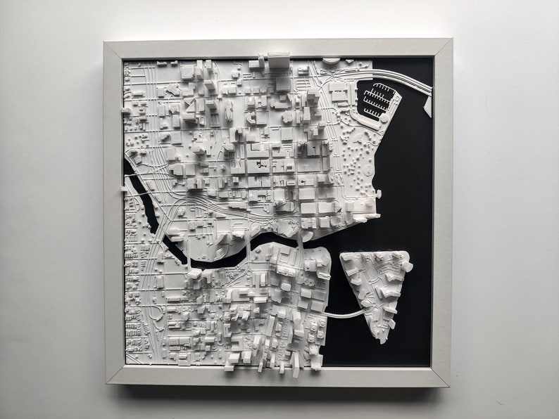 Puede incluir: Un modelo impreso en 3D de un horizonte de ciudad, en blanco, enmarcado en blanco y sobre un fondo negro. El modelo presenta edificios y calles detalladas, con un r&iacute;o que serpentea por la ciudad.
