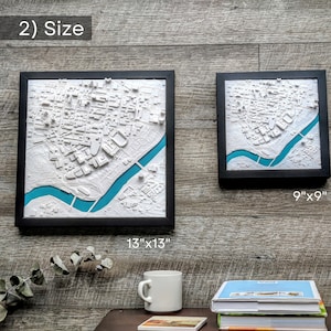 K&ouml;nnte beinhalten: Zwei gerahmte 3D-Stadtkarten, eine ist 13 Zoll mal 13 Zoll gro&szlig; und die andere 9 Zoll mal 9 Zoll. Beide Karten sind wei&szlig; mit einem t&uuml;rkisfarbenen Fluss und in Schwarz gerahmt.