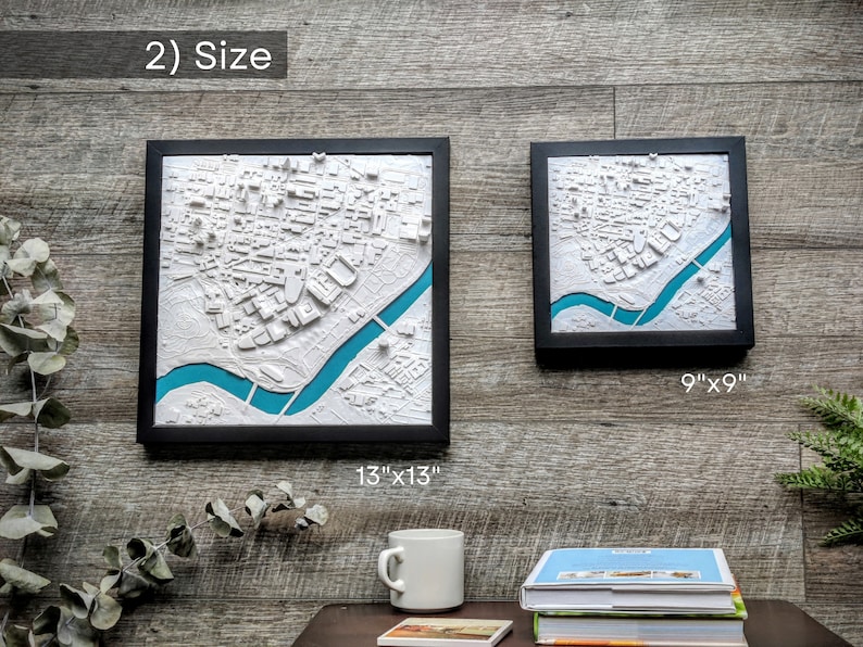 K&ouml;nnte beinhalten: Zwei gerahmte 3D-Stadtkarten, eine ist 13 Zoll mal 13 Zoll gro&szlig; und die andere 9 Zoll mal 9 Zoll. Beide Karten sind wei&szlig; mit einem t&uuml;rkisfarbenen Fluss und in Schwarz gerahmt.