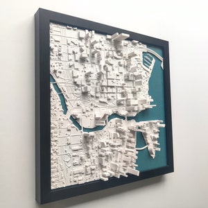 Puede incluir: Un modelo de ciudad en 3D enmarcado de una ciudad con un r&iacute;o que la atraviesa. El modelo es blanco y est&aacute; sobre un fondo azul verdoso. El marco es negro.