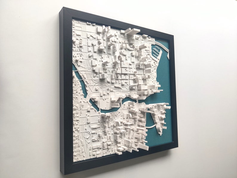 Puede incluir: Un modelo de ciudad en 3D enmarcado de una ciudad con un r&iacute;o que la atraviesa. El modelo es blanco y est&aacute; sobre un fondo azul verdoso. El marco es negro.