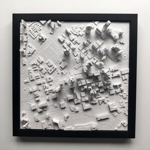 May include: A white 3D printed model of a city skyline in a black frame. The model is raised and shows the buildings in a city.
