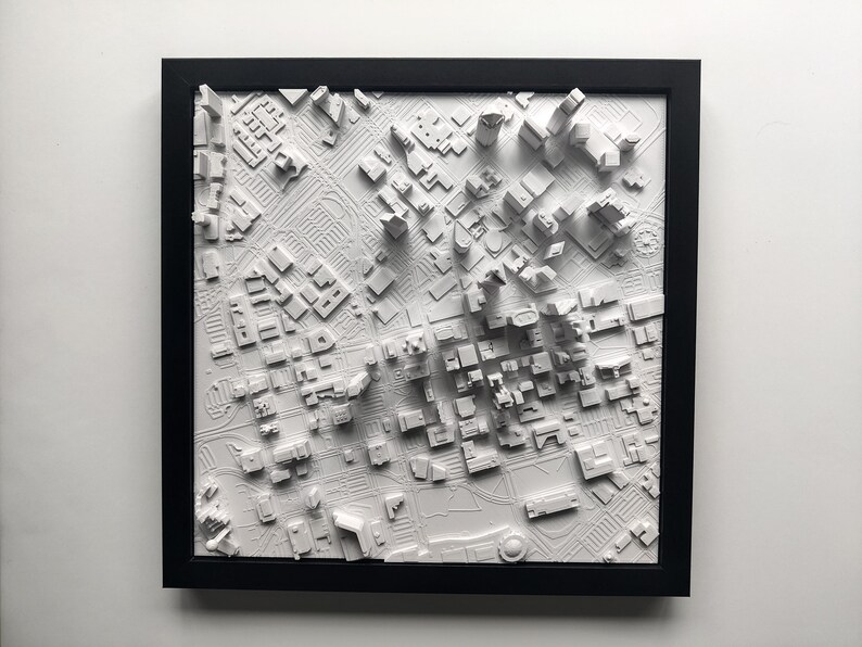 May include: A white 3D printed model of a city skyline in a black frame. The model is raised and shows the buildings in a city.