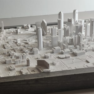 May include: A white 3D printed model of a city skyline, framed in a gray wood frame. The model features tall buildings and a detailed cityscape.