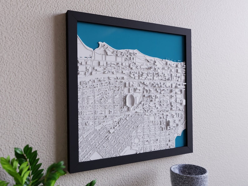 K&ouml;nnte beinhalten: Eine gerahmte 3D-Karte einer Stadt mit wei&szlig;en Geb&auml;uden und einem blauen Wassermerkmal. Die Karte ist in Schwarz gerahmt und h&auml;ngt an einer wei&szlig;en Wand.