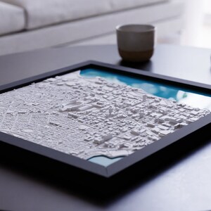 K&ouml;nnte beinhalten: Ein gerahmtes 3D-Modell einer Stadtlandschaft mit wei&szlig;en Geb&auml;uden und einem blauen Wassermerkmal. Der Rahmen ist schwarz und das Modell steht auf einem dunklen Tisch.