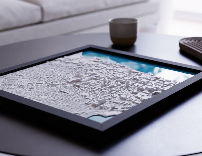 K&ouml;nnte beinhalten: Ein gerahmtes 3D-Modell einer Stadtlandschaft mit wei&szlig;en Geb&auml;uden und einem blauen Wassermerkmal. Der Rahmen ist schwarz und das Modell steht auf einem dunklen Tisch.