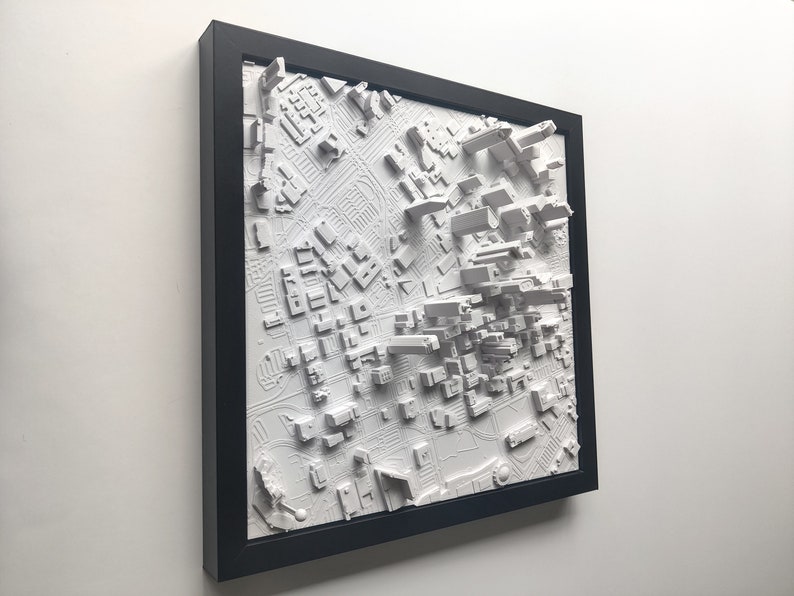 May include: A white 3D printed model of a city skyline in a black frame. The model is raised and textured to show the different buildings and streets.