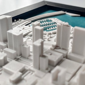 Puede incluir: Un modelo de ciudad en 3D impreso en blanco con una caracter&iacute;stica de agua azul. El modelo est&aacute; enmarcado y parece ser una ciudad en miniatura.