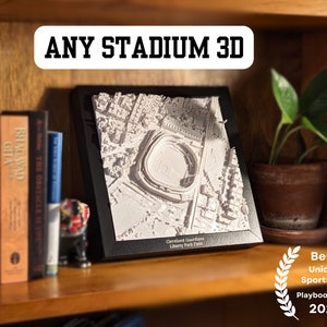 May include: A 3D model of a baseball stadium in a black frame, displayed on a wooden shelf with books and a potted plant. The model is white and shows the stadium's layout and surrounding area. The text "Cleveland Guardians Liberty Park Ewid" is visible below the model. The text "Best Unique Sports Gift Playbook Picks 2024" is visible below the shelf.