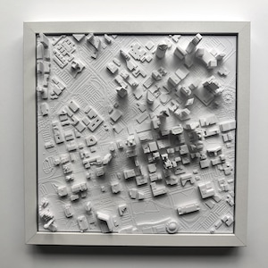 May include: A white 3D printed model of a city skyline, framed in a white frame. The model shows a detailed cityscape with buildings of varying heights.
