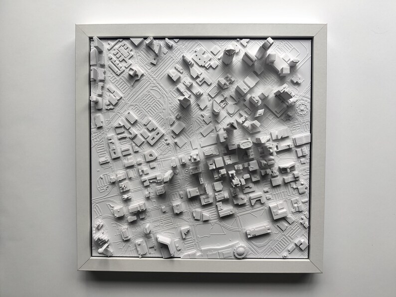 May include: A white 3D printed model of a city skyline, framed in a white frame. The model shows a detailed cityscape with buildings of varying heights.