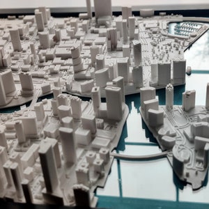 Puede incluir: Un modelo 3D blanco de un horizonte de ciudad, con edificios y carreteras, colocado en un marco de vidrio con un fondo azul.